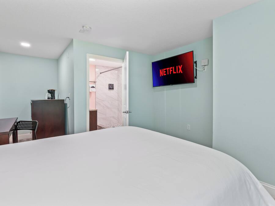 A hotel room with a bed, a desk, a chair, a TV mounted on the wall, and a door leading to a bathroom.