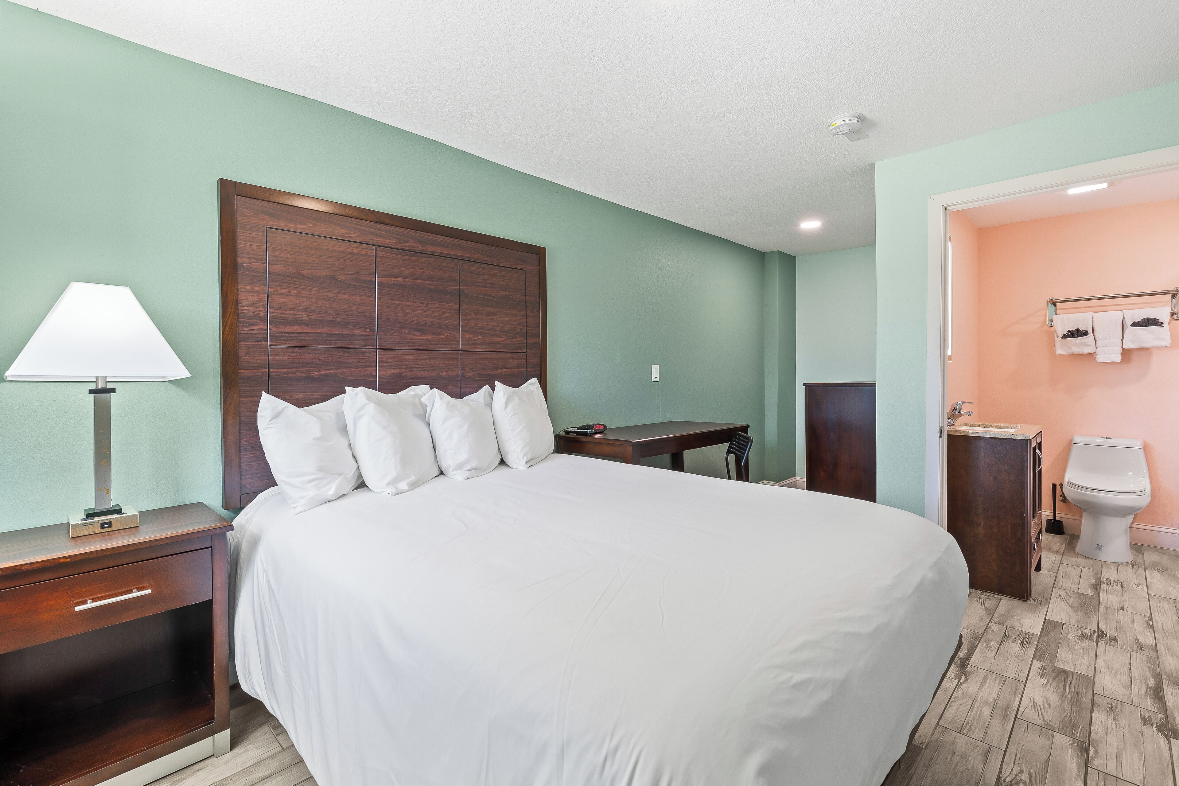 Hotelkamer met een groot bed, nachtkastje met lamp, houten hoofdbord, kleine tafel, en een badkamer met toilet en wastafel.