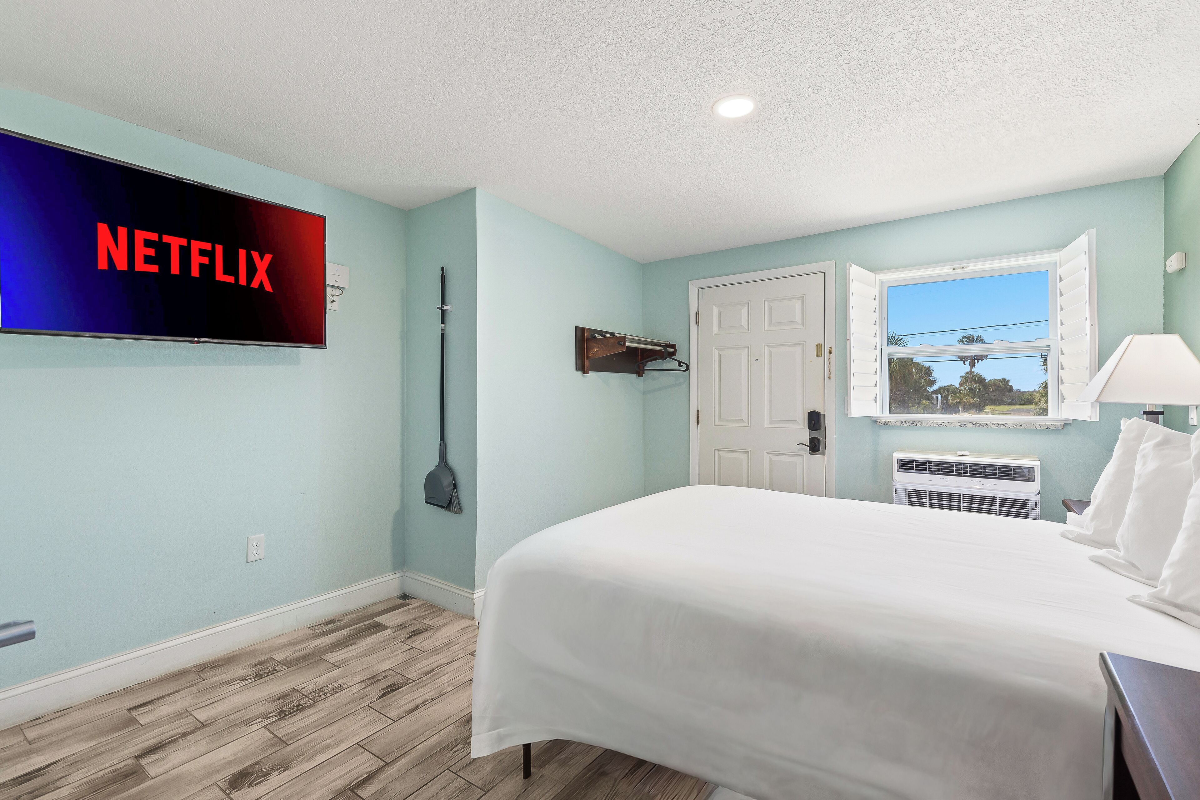 Un dormitorio con cama, televisor de pared mostrando 'NETFLIX', puerta, ventana con persianas y unidad de aire acondicionado.