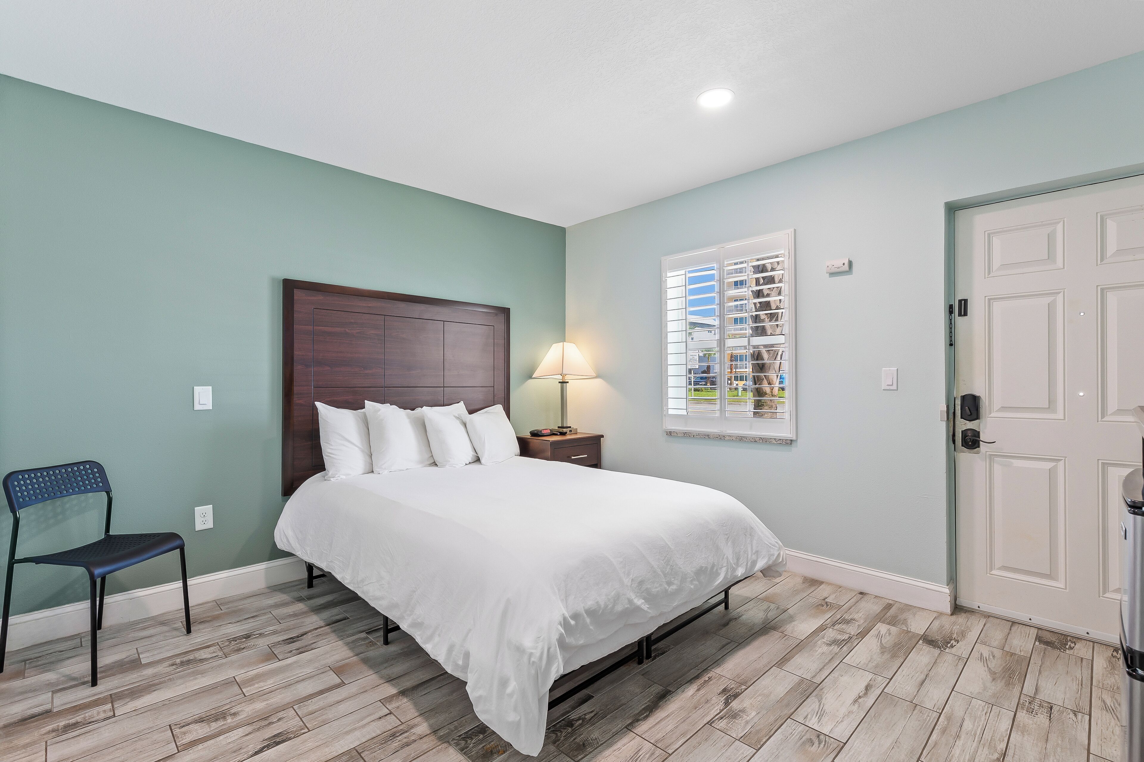 A bedroom with a bed, a nightstand, a lamp, a chair, and a window with blinds.