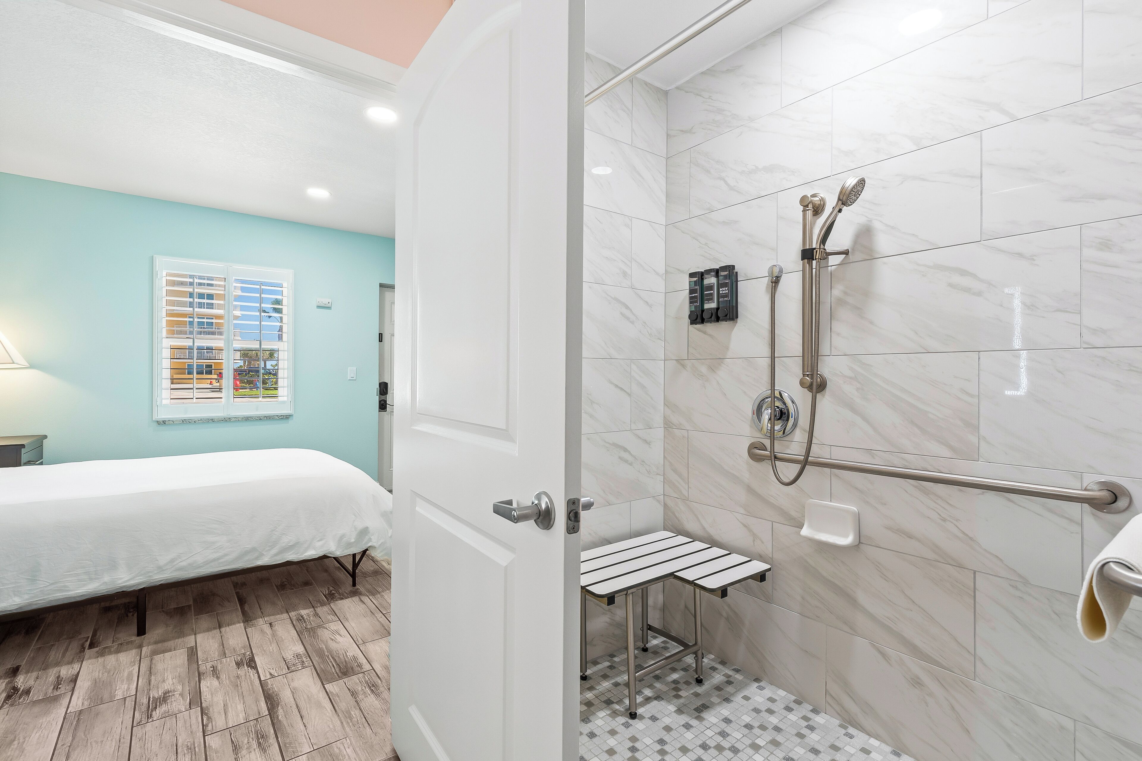 Habitación de hotel con cama, ducha con barra de apoyo y ventana con persianas.