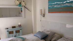 Ein Schlafzimmer mit einem Bett, Kissen, einem Nachttisch, einem Spiegel, einer Wandleuchte und einem Bild mit Strandmotiv.