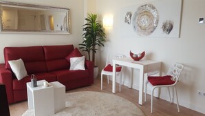 Ein Wohnzimmer mit einem roten Sofa, weißen Sesseln, einem kleinen Tisch, einer Topfpflanze und einem wandmontierten Spiegel.