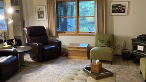 A living room with a brown leather chair, a green armchair, and a beige ottoman. There is a window with curtains, a lamp, and a framed picture on the wall. - Muskegon River Front Cabin  (Newaygo)