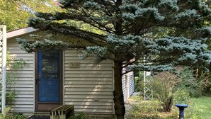 A small, single-story house with a blue door and a number '7168' on the side. - Muskegon River Front Cabin  (Newaygo)