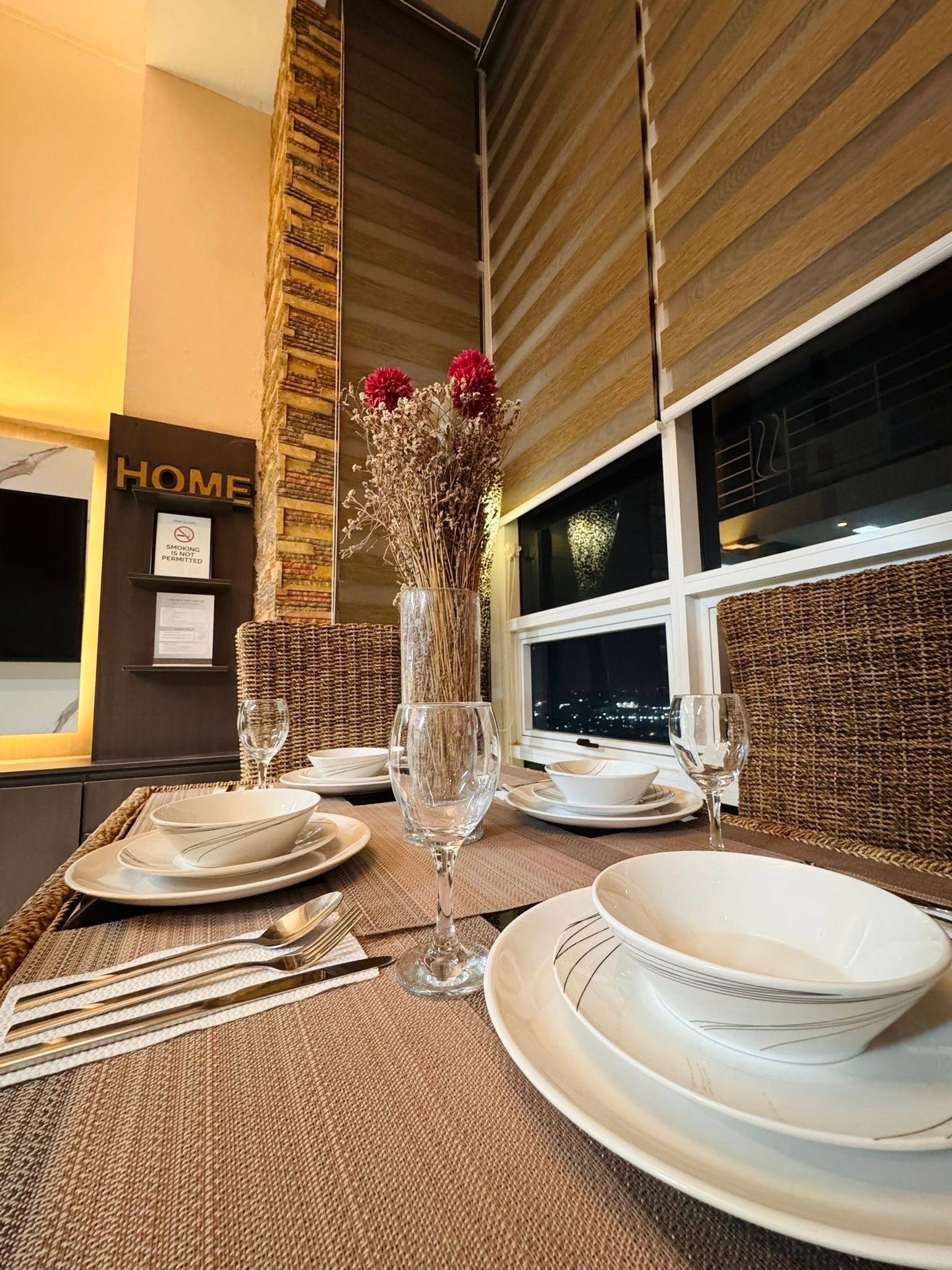 A dining table set with white plates, silverware, and wine glasses, with a vase of dried flowers and a 'HOME' sign on the wall.