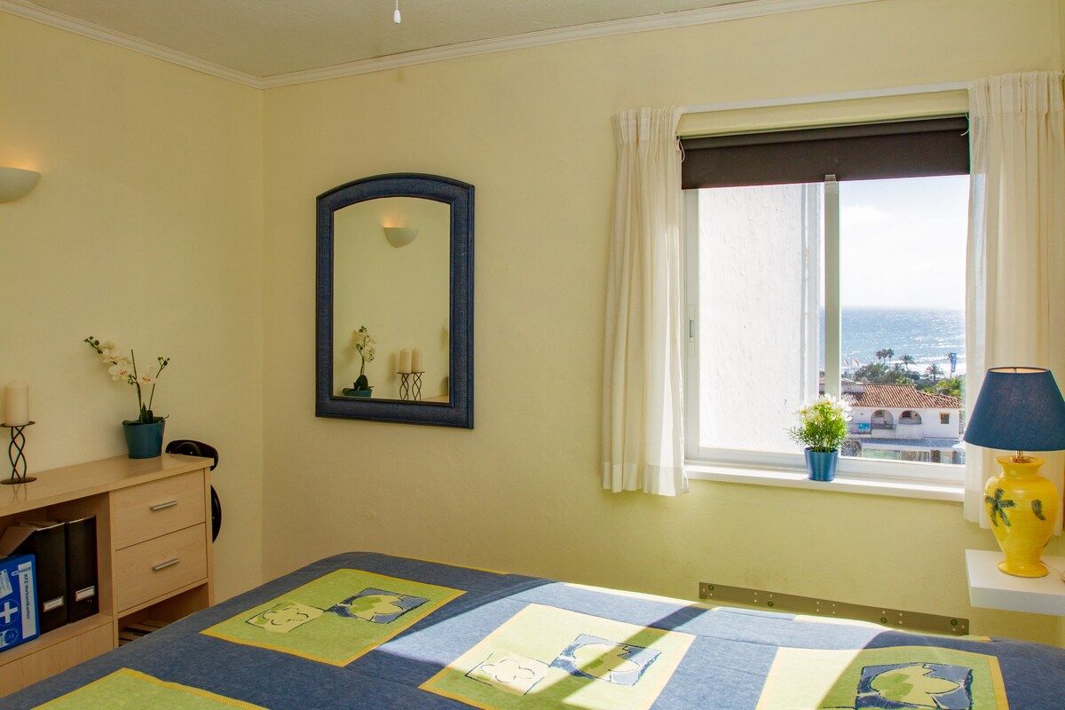 Ein Schlafzimmer mit einem Bett, einem Schminktisch, einem Spiegel, einem Fenster mit Vorhängen und Blick aufs Meer.