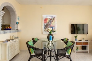 A dining area with a round glass table, wicker chairs, a vase with flowers, a framed abstract painting, a television, and a kitchen area with a brick archway. - Riviera Playa | Charming Apartment with Sea Views, Terrace & Pools (Mijas Costa)