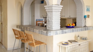 A kitchen with a stone countertop, two wicker bar stools, and a white cabinet with a blue bowl of fruit. - Riviera Playa - Costa del Sol - Charming apartment with Sea View (Mijas Costa)
