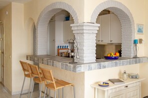 Une cuisine dotée d’un plan de travail en pierre, de deux tabourets de bar en osier et d’un meuble blanc sur lequel est posé un saladier bleu.