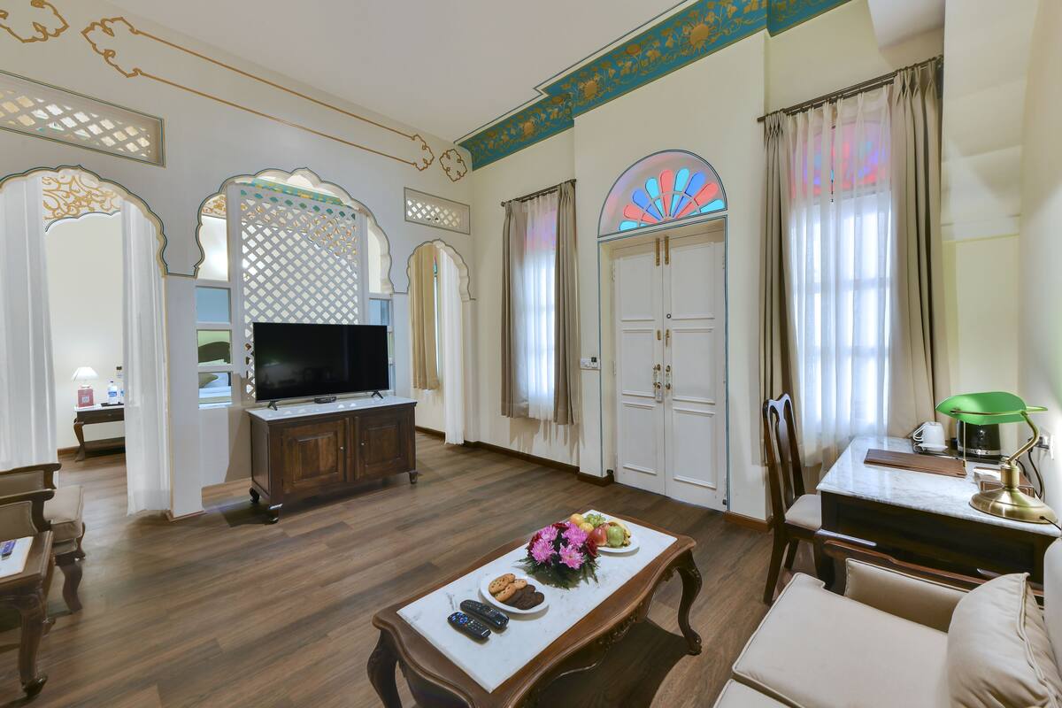 A spacious living room with a wooden floor, a television on a wooden cabinet, a dining table with a vase of flowers, and a coffee table with remote controls and snacks.