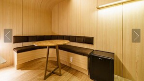 Un banco moderno de madera curvada con una pequeña mesa redonda y un electrodoméstico negro, probablemente un calentador, en una habitación con paneles de madera y flechas indicativas en las paredes.