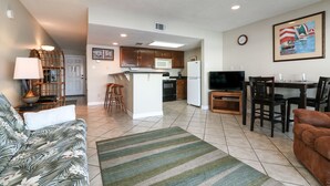 A modern living room with a sofa, a coffee table, and a striped rug. There is a kitchen area with a microwave, oven, and refrigerator. A dining table with chairs is visible, and a painting hangs on the wall. - 1 Bedroom with Private Waterfront Balcony (Willis)