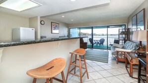 A kitchen with a refrigerator, wooden bar stools, and a dining area with a sofa and a lamp. - 1 Bedroom with Private Waterfront Balcony (Willis)