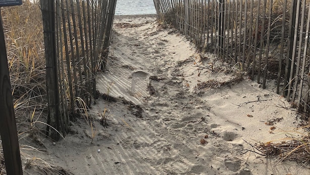 Ein Sandweg am Strand mit hölzernen Pfosten und einem Hinweisschild.