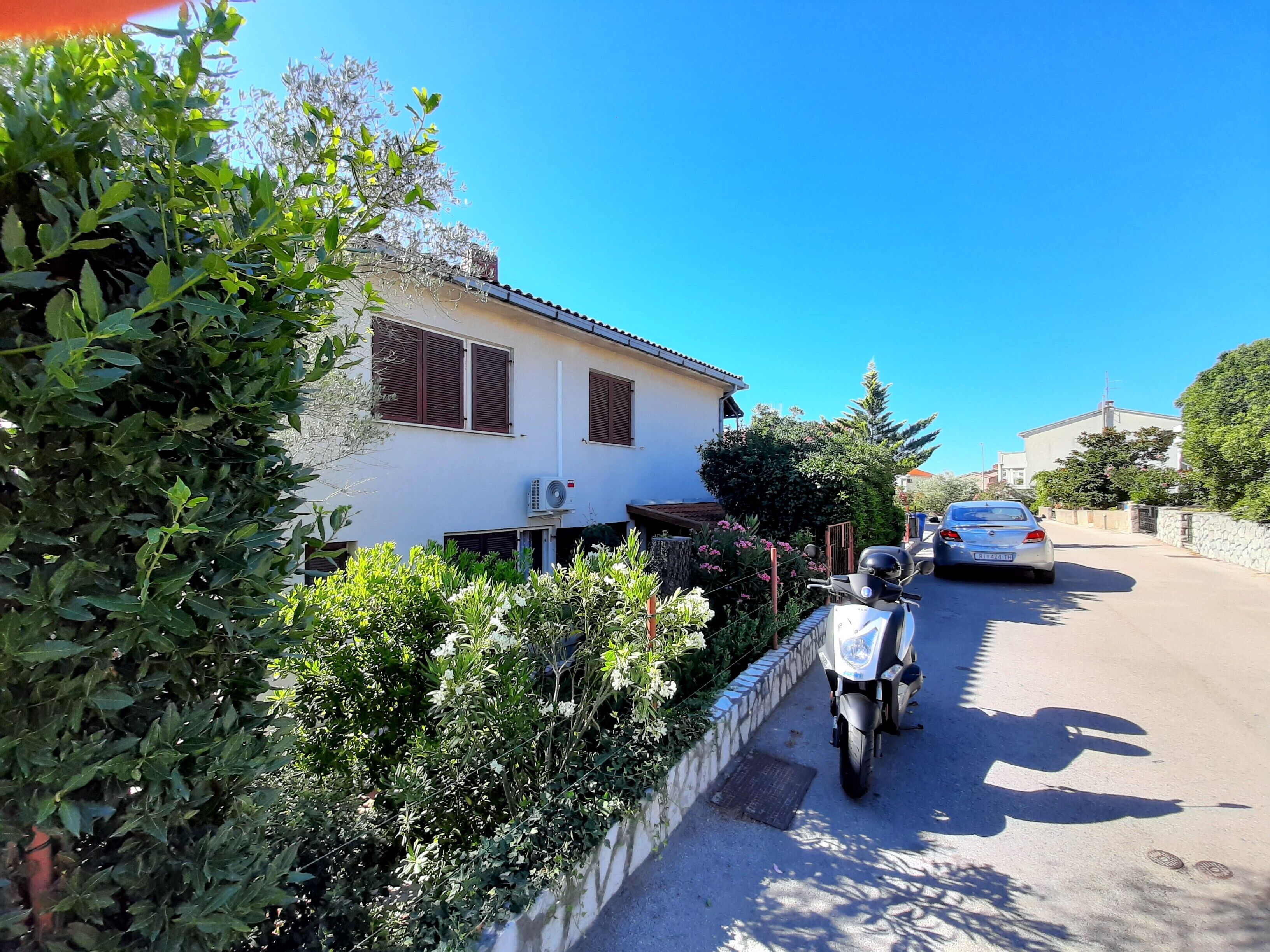 Una calle residencial con una casa blanca, un scooter estacionado y un coche.