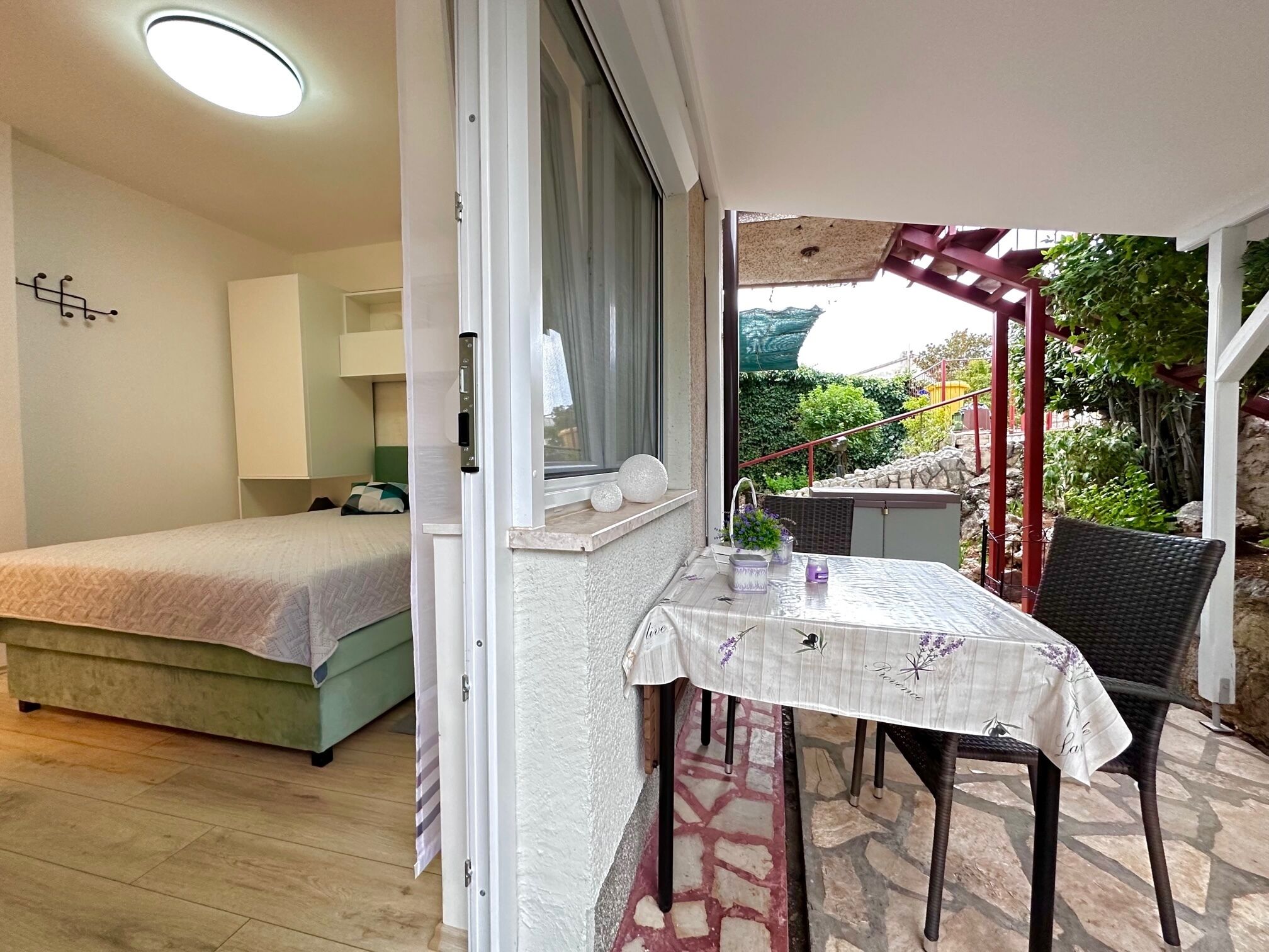A bedroom with a bed and a nightstand, a balcony with a table and chair, and a view of greenery.