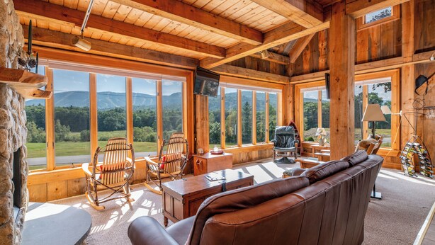 A cozy wooden cabin interior with a large window offering a scenic mountain view, a leather sofa, and rustic decor. - Mountain Lodge Retreat (Newry)