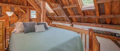 Ein Schlafzimmer im Landhausstil mit Schlafgalerie, geneigter Decke, einem Bett mit grüner Tagesdecke und einem Fenster mit Blick ins Grüne.