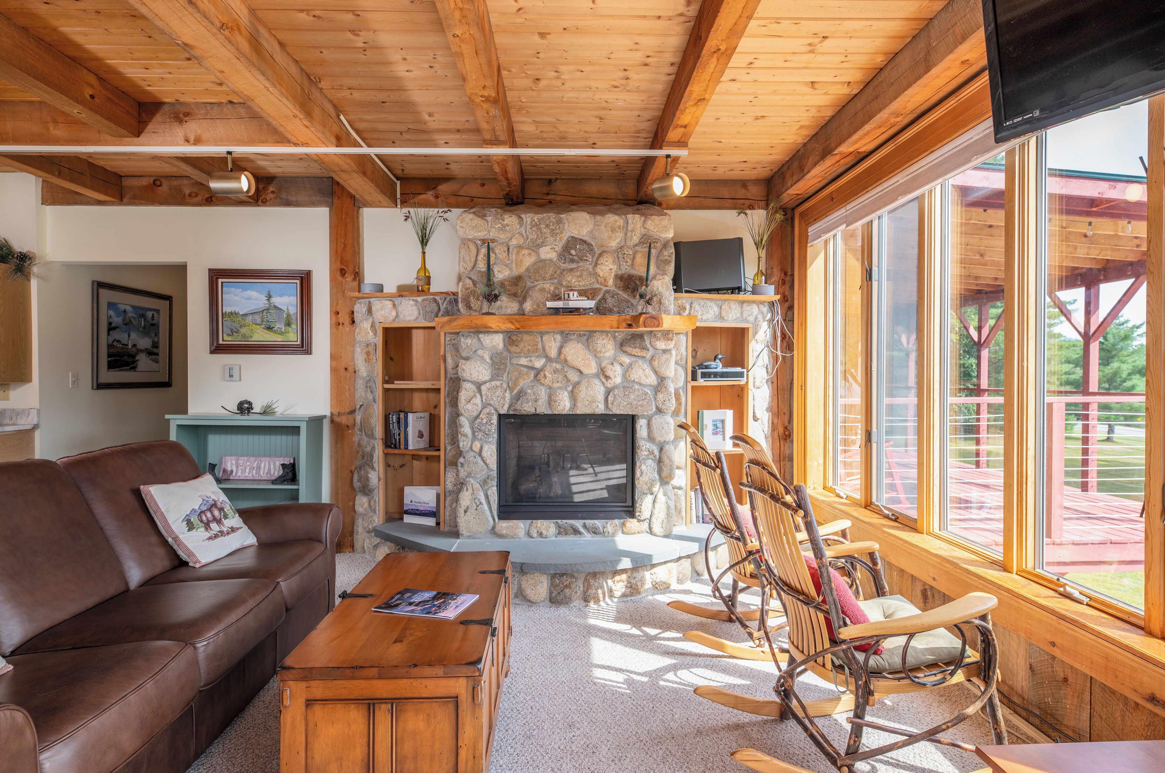 A cozy living room with a stone fireplace, wooden ceiling beams, a brown leather sofa, and a rocking chair.
