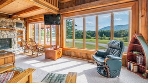 A cozy living room with a stone fireplace, wooden furniture, and large windows offering a view of a green landscape. - Mountain Lodge Retreat (Newry)