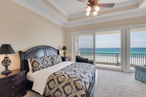 A bedroom with a large bed, two bedside tables with lamps, a ceiling fan, and a view of the beach through a sliding glass door. - Tuscany Sunrise - A Luxury Retreat for Discerning Guests (Navarre)