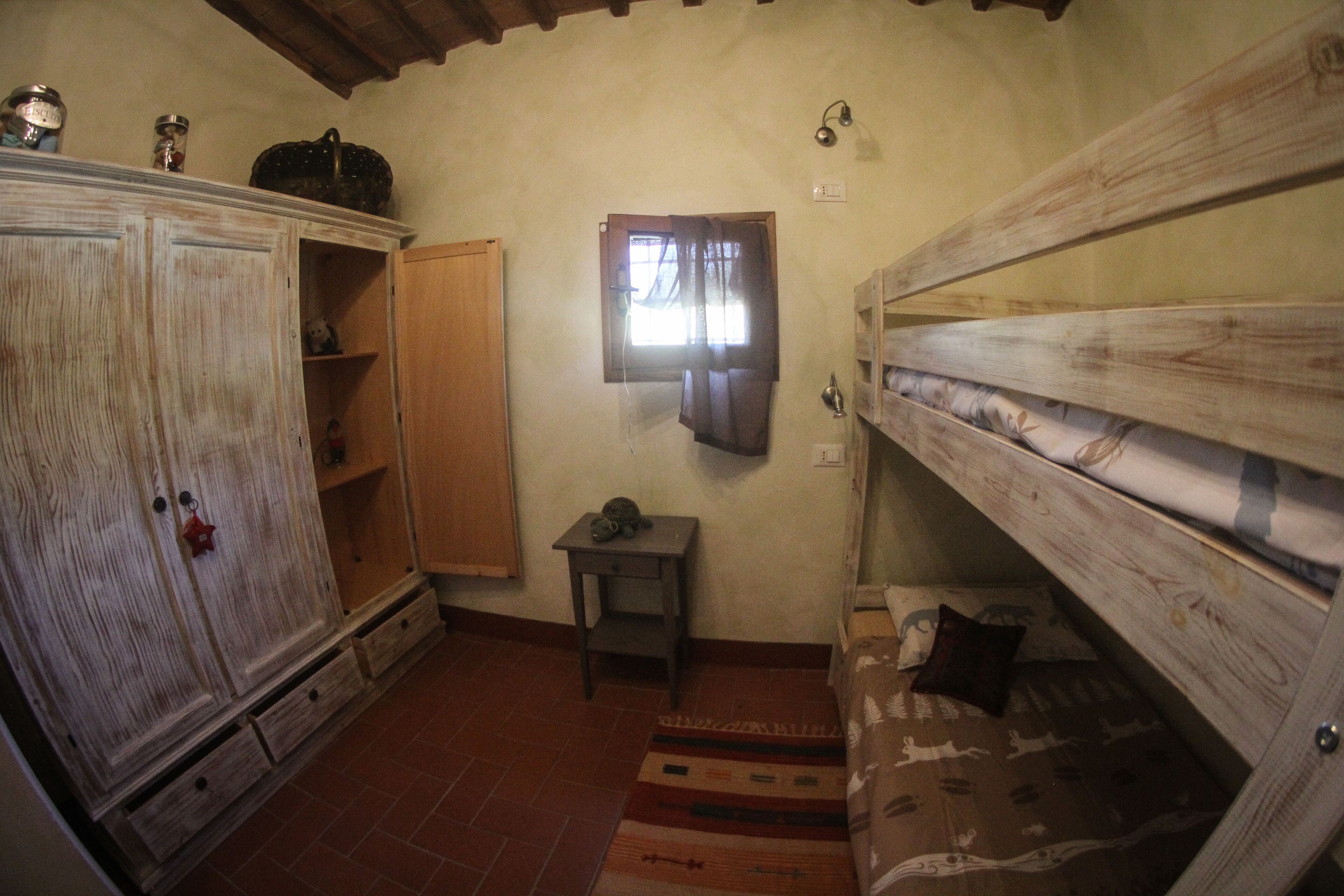 Ein kleiner, rustikaler Raum mit einem Holzbett, einem Schrank aus Holz, einem kleinen Tisch und einem Fenster mit Vorhängen.