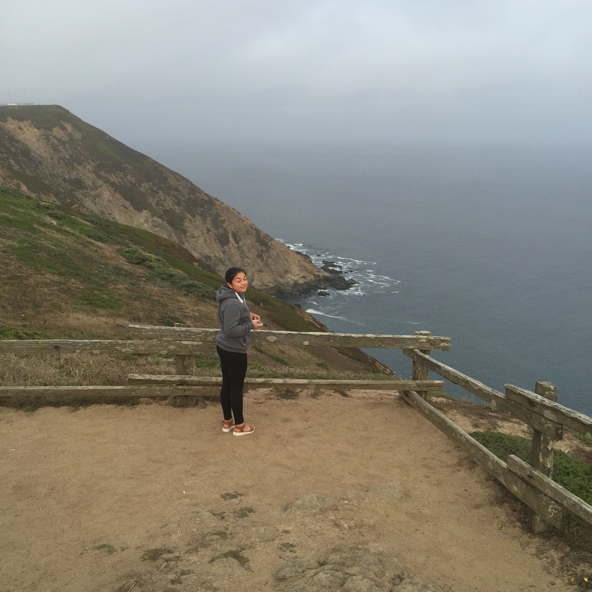 A person standing on a dirt path with a wooden railing, overlooking a coastal cliff and ocean.