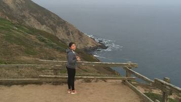 A person standing on a dirt path with a wooden railing, overlooking a coastal cliff and ocean.
