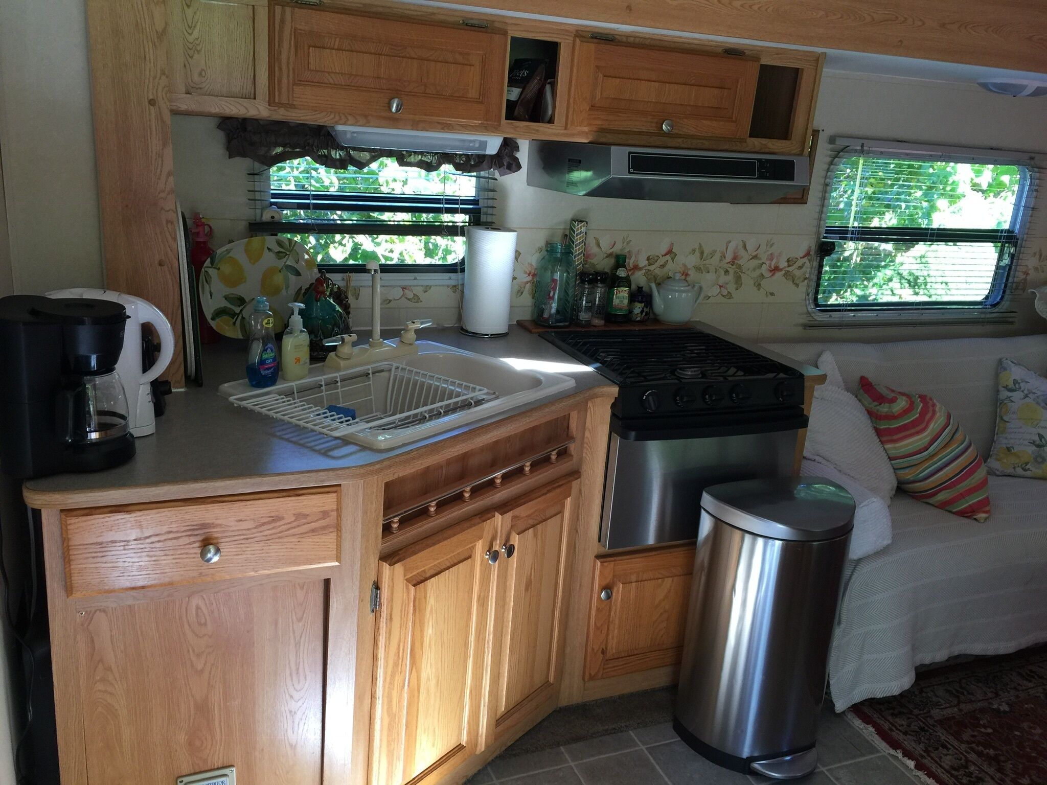 A compact kitchen with wooden cabinets, a stainless steel stove, a sink with a dish rack, and a trash can.