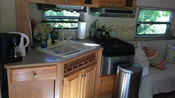 A compact kitchen with wooden cabinets, a stainless steel stove, a sink with a dish rack, and a trash can.