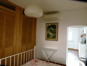Ein Zimmer mit Holzwänden, einer weißen Klimaanlage, einem Baumgemälde, einem weißen Klappstuhl und einem Blick auf einen Küchenbereich.