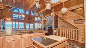 A wooden cabin interior with a kitchen, large windows, and a stone fireplace. - A View To Remember: MTN VIEWS! Luxury, Hot Tub, Wifi, Fireplace, Games and more (Sevierville)