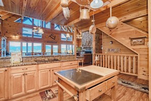Interior de una cabaña de madera con cocina, ventanas amplias y chimenea de piedra.