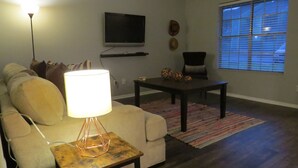Un salon avec un canapé, une table basse, une lampe, un téléviseur et une fenêtre avec des stores.