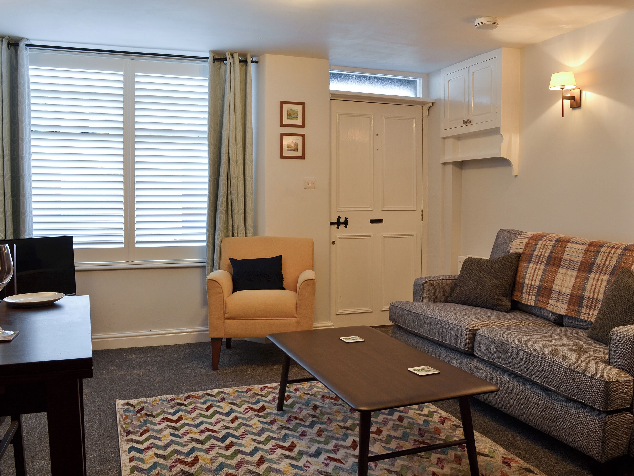 Un salon comprenant un canapé, un fauteuil, une table basse et une table à manger avec des chaises. Il y a une fenêtre avec des stores, une porte et une lampe fixée au mur.