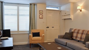 Een woonkamer met een bank, fauteuil, salontafel en een eettafel met stoelen. Er is een raam met jaloezieën, een deur en een wandlamp.