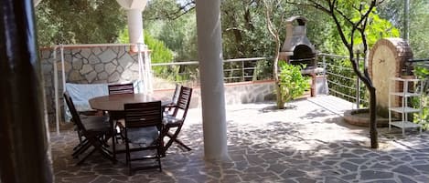 A covered patio with stone flooring, a table with chairs, and a brick oven.