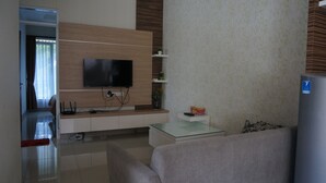 A modern living room with a flat-screen TV mounted on a wooden panel wall, a glass coffee table, a sofa, and a small shelf with decorative items. - Ouma Guest House (Bandung)