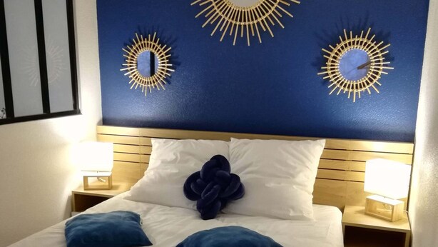 Un lit bien fait, avec du linge de lit blanc et des coussins bleus, deux miroirs décoratifs en forme de soleil fixés au mur, et des lampes de chevet posées sur des tables de nuit en bois.