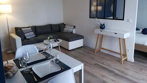 Une pièce moderne et bien éclairée, avec un canapé gris, une table à manger dressée avec des assiettes et des verres, une petite table d’appoint et un bureau blanc agrémenté d’un miroir.