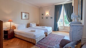 A hotel room with two beds, a wooden floor, a patterned rug, a nightstand, a lamp, and a view of the outdoors through a window with curtains. - ST GERMAIN, BALCONY, VIEW, 2 BATHS, 30–60 days ahead? Private offer. BEV-VBs (Paris)