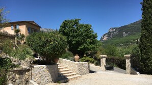 Un edificio de paredes de piedra con un jardín, un árbol grande y una montaña al fondo.