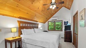 A bedroom with a wooden ceiling, a bed, a nightstand, a lamp, a TV, and a window with a view of greenery. - Treetop Views, a Stone Fireplace, and Mountain Fresh Market Around the Corner | Satulah Chalet (Highlands)
