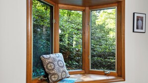 A bay window with a cushion and a book on the sill, overlooking a lush garden. - Treetop Views, a Stone Fireplace, and Mountain Fresh Market Around the Corner | Satulah Chalet (Highlands)