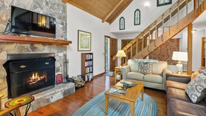 A cozy living room with a stone fireplace, wooden beams, a staircase, and a comfortable seating area.