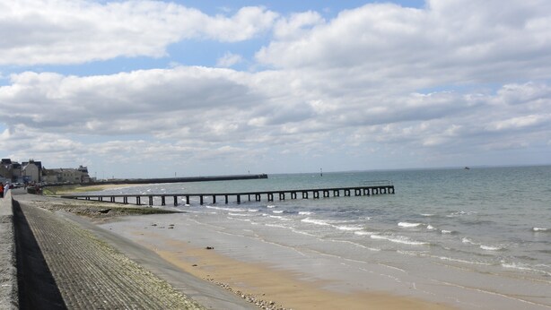 Une plage avec une jetée en bois qui s’avance dans l’eau, une rive sablonneuse et des bâtiments en arrière-plan.
