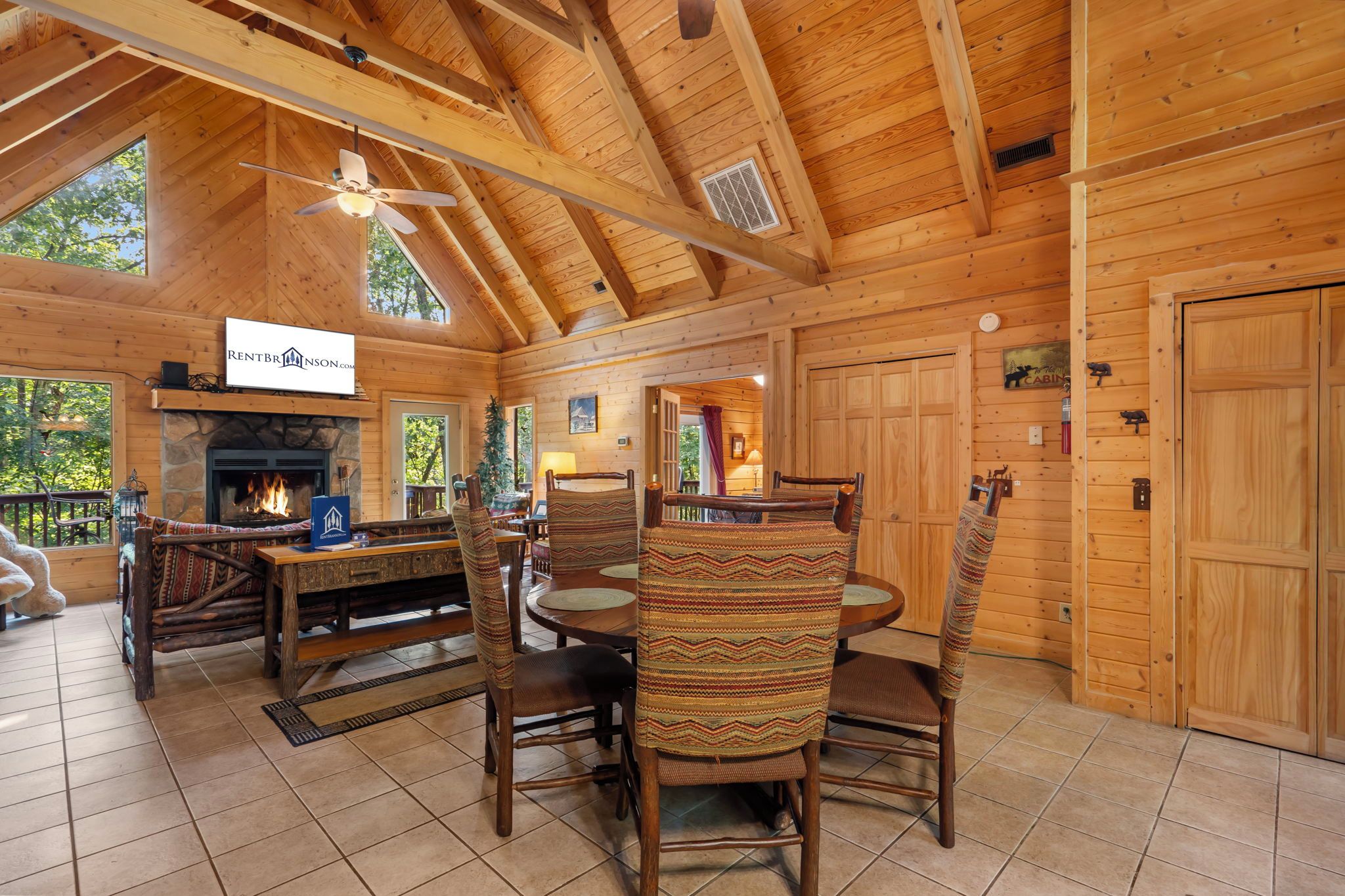 A log cabin interior with a fireplace, dining area, and a ceiling fan.