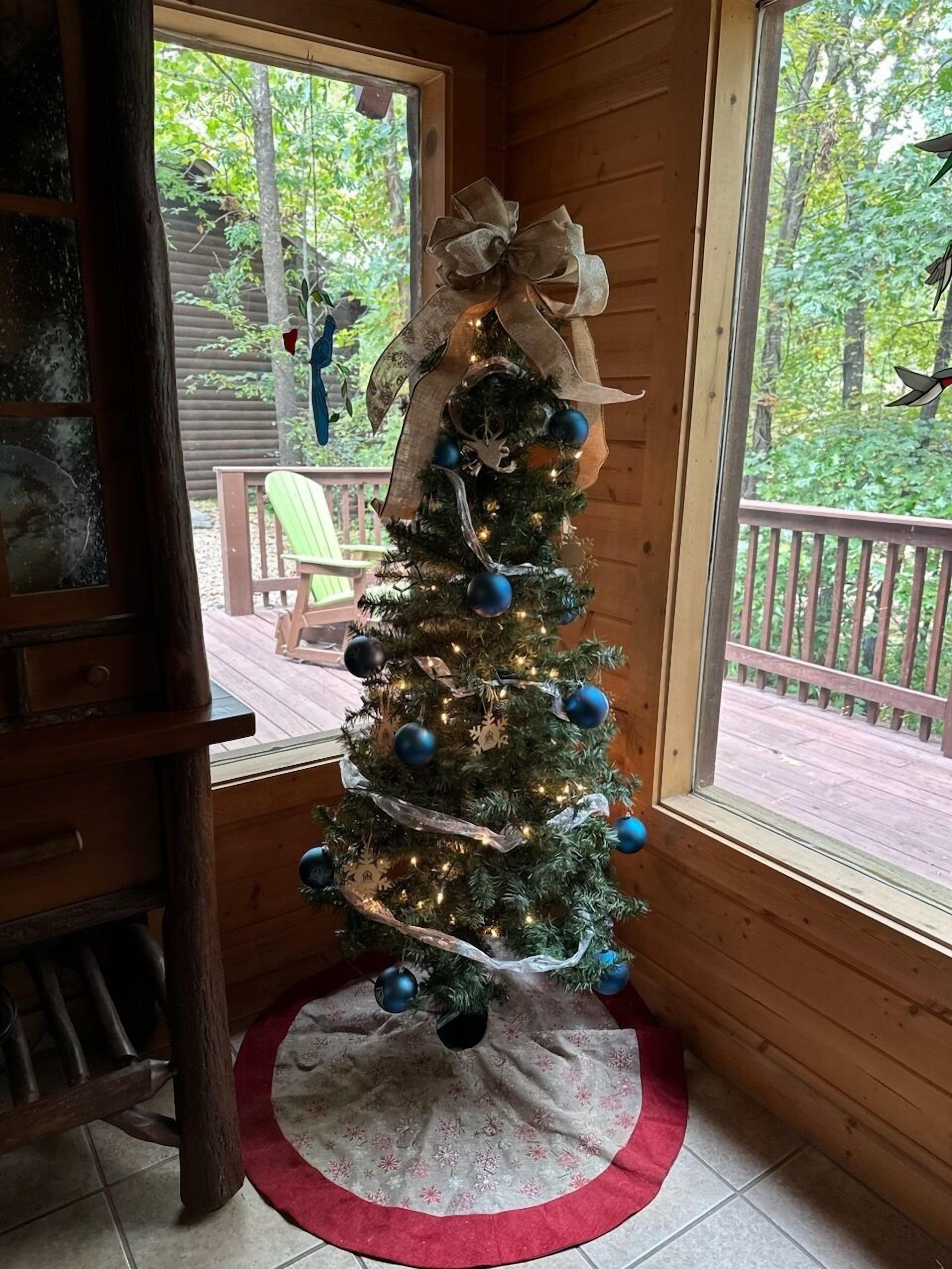 Ein mit blauen Ornamenten und einer großen Schleife geschmückter Weihnachtsbaum, platziert in einer Holzhütte mit Blick auf eine Terrasse und Bäume.