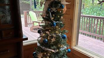 Ein mit blauen Ornamenten und einer großen Schleife geschmückter Weihnachtsbaum, platziert in einer Holzhütte mit Blick auf eine Terrasse und Bäume.